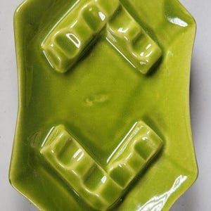 May include: A green ceramic ashtray with four indented cigarette rests. The ashtray is shaped like a diamond with rounded corners.