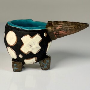 May include: A small ceramic vessel with a black exterior featuring white circular and cross-shaped patterns. The interior is glazed in bright turquoise. It has three short, textured brown legs and a prominent, pointed brown handle extending outward.