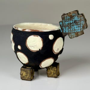 May include: A small, round, black ceramic cup featuring white polka dots and a textured, light-colored interior. The cup stands on three small, square, gold-toned feet and has a single, square, blue-and-brown textured handle extending outward.