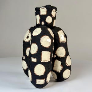 May include: A sculptural ceramic vessel with a matte black finish, featuring an irregular, rounded form. The surface is decorated with numerous hand-applied, off-white, glossy geometric shapes, including various circles and squares.