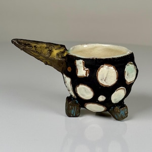 May include: A small, hand-built ceramic cup with a black exterior featuring white circular and rectangular spots. The cup has a long, pointed handle extending from one side and stands on three short, blocky feet with blue accents on the base.