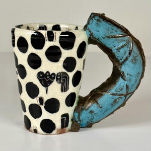 May include: A ceramic mug with a cream-colored base featuring black polka dots and small hand-drawn floral motifs. The mug has a large, textured, C-shaped handle finished in a bright turquoise blue glaze with dark brown, rustic, metallic-looking edges.