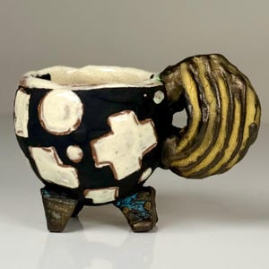May include: A small ceramic cup with a matte black exterior featuring raised white geometric shapes including crosses and circles. The cup has three short, textured feet and a large, prominent handle with alternating yellow and dark brown stripes.