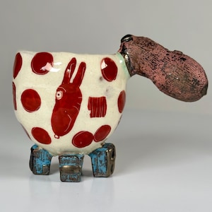 May include: A ceramic cup with a cream-colored base featuring red polka dots and a red rabbit illustration. The cup has three textured blue feet and a large, protruding, textured pink handle extending from the side, resembling a stylized animal head.