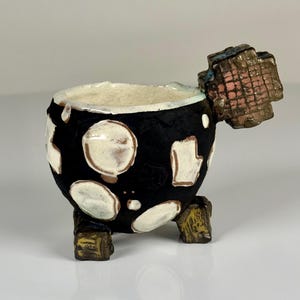 May include: A small, round, black ceramic cup with an uneven, textured surface featuring irregular white patches. It stands on three short, blocky, metallic-toned feet and has a prominent, square, grid-patterned handle extending from the side.