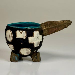 May include: A small, round, black ceramic cup with a bright turquoise interior. The exterior features irregular white shapes, including a cross. It has a textured, bronze-colored handle extending to the right and stands on three short, matching bronze-colored feet.