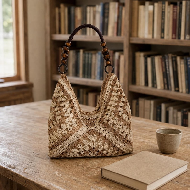 Puede incluir: Un bolso de ganchillo en tonos beige y marr&oacute;n con un patr&oacute;n de cuadrados granny y un asa marr&oacute;n oscuro decorada con cuentas de madera redondas. El bolso tiene un dise&ntilde;o estructurado de apertura superior con una silueta triangular.