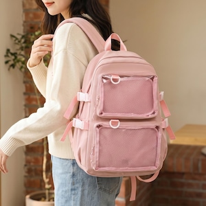 Puede incluir: Una mochila rosa con dos bolsillos delanteros con diseño de malla. La mochila tiene correas ajustables y hebillas blancas. La bolsa está hecha de un material suave y tiene un asa superior.