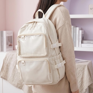 Puede incluir: Mochila de color crema con múltiples bolsillos y correas ajustables. Cuenta con dos bolsillos frontales transparentes y un bolsillo de malla. La mochila tiene un asa superior y está diseñada para llevar artículos escolares o de viaje.