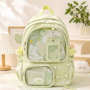 Puede incluir: Una mochila verde claro con un bolsillo frontal transparente que muestra pegatinas en forma de corazón y un tarjetero a juego. La mochila tiene bolsillos laterales, un asa superior y detalles decorativos. El diseño es suave y en tonos pastel.