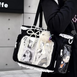 May include: Black tote bag with white lace trim and a clear front pocket displaying manga art and a plushie. The bag has a shoulder strap and is decorated with a bow, a fluffy charm, and a pearl necklace.