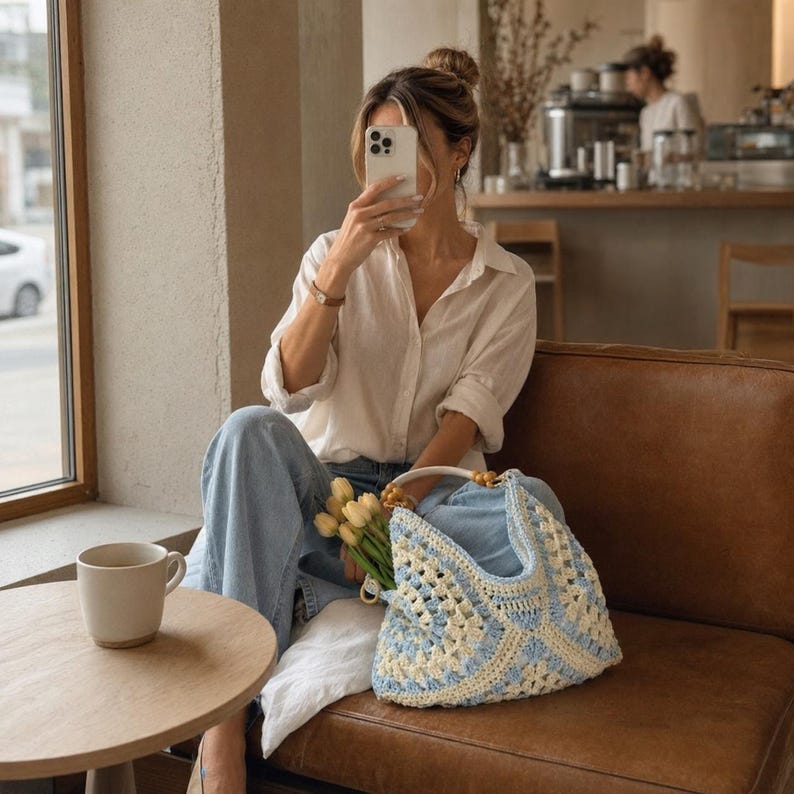 Puede incluir: Una camisa blanca de botones, pantalones vaqueros azul claro y un bolso de ganchillo en tonos azul claro y crema con un asa de cuentas de madera. El bolso contiene un peque&ntilde;o ramo de tulipanes amarillos. Una taza de cer&aacute;mica blanca sobre una mesa.