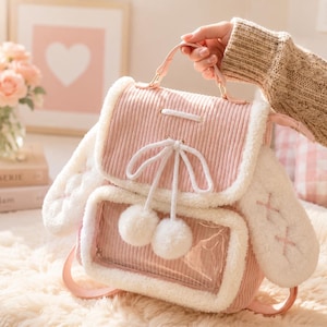 Puede incluir: Una mochila rosa de pana con ribete blanco y esponjoso y detalles de orejas de conejo. La bolsa tiene un bolsillo frontal transparente, un asa superior y un lazo blanco con dos pompones. La bolsa está sujeta por una persona que lleva un suéter beige.
