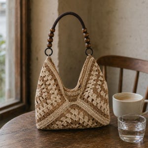 Puede incluir: Un bolso de ganchillo con un patr&oacute;n de cuadrados granny en beige y marr&oacute;n. El bolso tiene una abertura superior curva y un asa &uacute;nica adornada con cuentas de madera marr&oacute;n oscuro a cada lado, unida al cuerpo mediante anillos circulares.