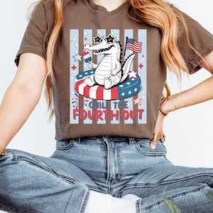 Könnte beinhalten: Braunes T-Shirt mit einer Grafik eines Alligators mit sternförmiger Sonnenbrille und Eis am Stiel, der auf einem patriotischen Schwimmring schwimmt. Der Text "Chill the Fourth Out" steht darunter. Enthält eine kleine amerikanische Flagge.