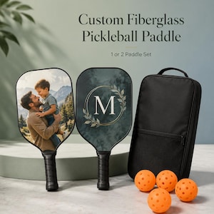 Könnte beinhalten: Zwei personalisierte Pickleball-Schläger aus Fiberglas mit schwarzen Griffen. Ein Schläger zeigt ein Foto einer Person mit Kind, der andere ein M in einem goldenen Kranz. Enthält eine schwarze Tragetasche und vier orangefarbene Pickleballs.
