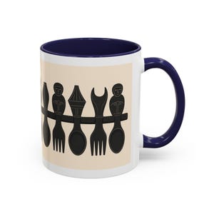 May include: White ceramic mug with a navy blue interior and handle. The mug features a beige band with a black graphic of stylized utensils and figures. The design includes spoons, forks, and a decorative element.