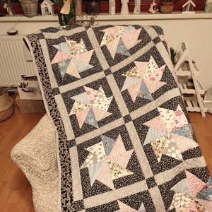 Puede incluir: Un edredón hecho a mano con un diseño de estrella. El edredón presenta un borde floral negro y gris, con un diseño central de cuadrados negros alternos y bloques en forma de estrella en patrones rosas, azules y florales. El edredón está sobre una silla.