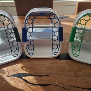 Peut inclure: Trois mangeoires pour animaux avec des bases blanches et des couvercles arqués transparents. Chaque mangeoire présente un cadre à motif nid d'abeille de couleur noire, bleue ou verte. Les unités ont des bols internes pour la nourriture.