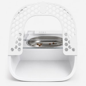 Op de afbeelding: Een witte voerbak voor huisdieren met een boogvormig frame met honingraatpatroon en een doorzichtige plastic kap. De unit heeft een centrale ovale roestvrijstalen kom met enkele brokjes droogvoer. Voorzien van een brede basis en handvat.