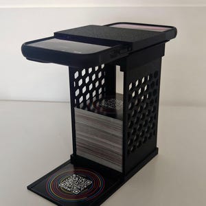 Könnte beinhalten: Ein schwarzer 3D-gedruckter Ständer mit wabenförmiger Seitenstruktur, der für einen Stapel quadratischer Karten ausgelegt ist. Ein Smartphone liegt horizontal auf der oberen Plattform, während eine einzelne Karte mit einem bunten QR-Code-Grafik auf der Basis liegt.