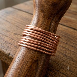 Może przedstawiać: Stos dziesięciu cienkich, gładkich, metalicznych bransoletek w kolorze miedzi, owiniętych wokół polerowanego drewnianego stojaka. Bransoletki mają prosty, zaokrąglony kształt drutu z jednolitą, odblaskową powierzchnią w ciepłym, czerwonobrązowym odcieniu.