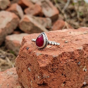 Op de afbeelding: Een zilveren ring met een peervormige, gefacetteerde dieprode edelsteen in een zetting. De ringband is opgebouwd uit een reeks kleine, verbonden zilveren bolletjes en de zetting van de steen is omlijst met een gedraaid zilveren draadje.