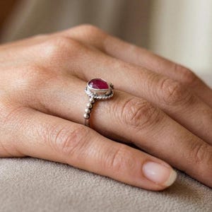 Op de afbeelding: Een zilveren ring met een peervormige, gefacetteerde dieproze edelsteen in een zetting. De ringband is ontworpen met een getextureerd, kralenachtig patroon dat om de vinger loopt en een gedetailleerde metalen afwerking rond de steen geeft.