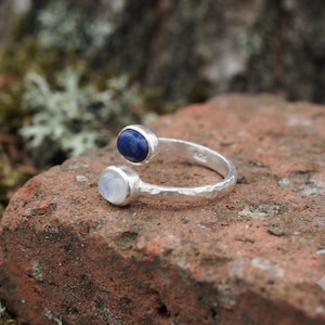 Peut inclure: Une bague ouverte en argent avec une bande à la texture martelée. Une extrémité est ornée d'une pierre ronde et lisse en lapis-lazuli bleu profond, tandis que l'autre extrémité porte un cabochon de pierre de lune rond, translucide et irisé.