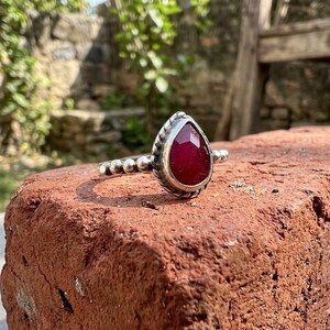 Op de afbeelding: Een zilveren ring met een peervormige, gefacetteerde dieprode edelsteen in een textuurrijke metalen zetting. De ringband bestaat uit een reeks kleine, uniforme zilveren bolletjes die vanaf de centrale zetting uitlopen in een kralenpatroon.