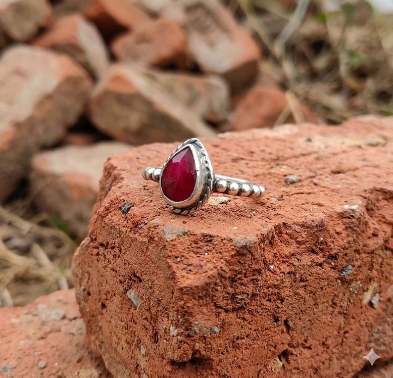 Op de afbeelding: Een zilveren ring met een peervormige, gefacetteerde dieprode edelsteen in een zetting. De ringband is opgebouwd uit een reeks kleine, verbonden zilveren bolletjes en de zetting van de steen is omlijst met een gedraaid zilveren draadje.