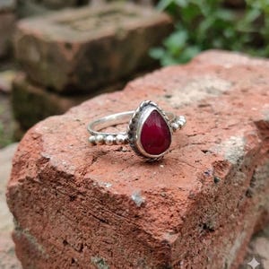 Può includere: Un anello in argento con una pietra preziosa rosso scuro a forma di goccia, incastonata in una lunetta di metallo intrecciato. La fascia è decorata con una serie di piccole perline d'argento rotonde su entrambi i lati della pietra centrale.