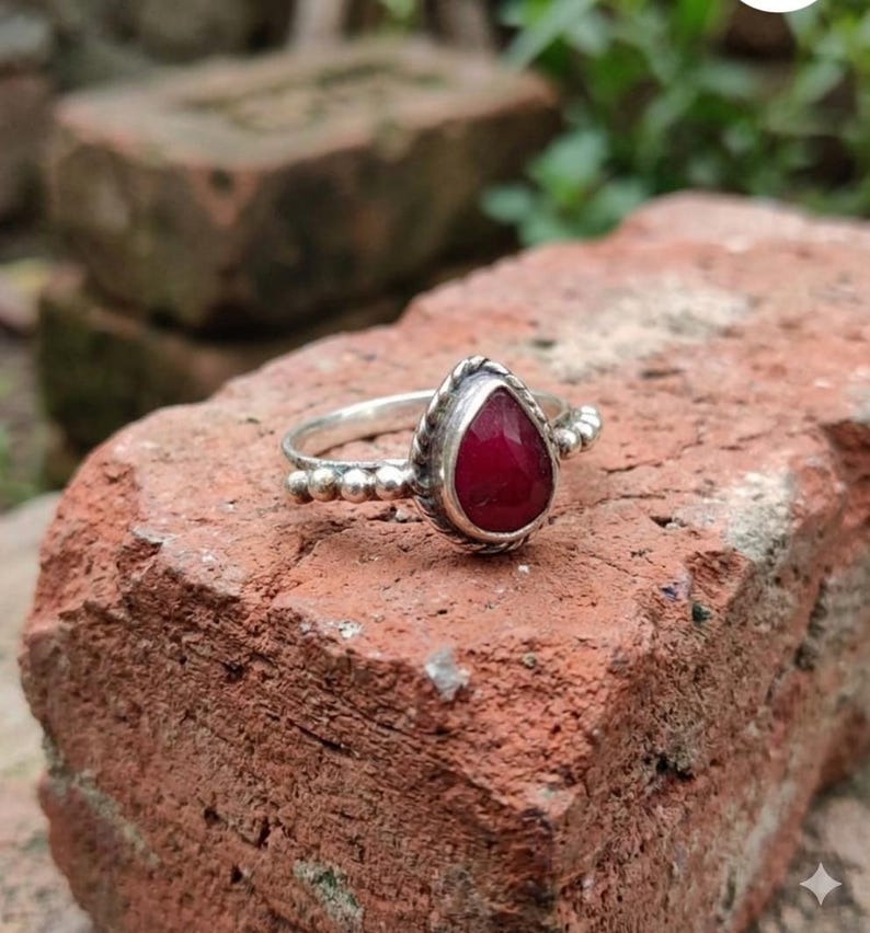 Op de afbeelding: Een zilveren ring met een druppelvormige, dieprode edelsteen gezet in een gedraaide metalen zetting. De ringband is aan beide zijden van de centrale steen versierd met een reeks kleine, ronde zilveren kraaltjes, wat zorgt voor een gedetailleerd uiterlijk.