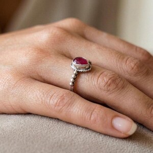 Op de afbeelding: Een zilveren ring met een peervormige, donkerroze edelsteen in een zetting. De ringband is opgebouwd uit een reeks kleine, bolvormige metalen kralen, die uitlopen in een gedraaid draaddetail rondom de centrale zetting van de edelsteen.