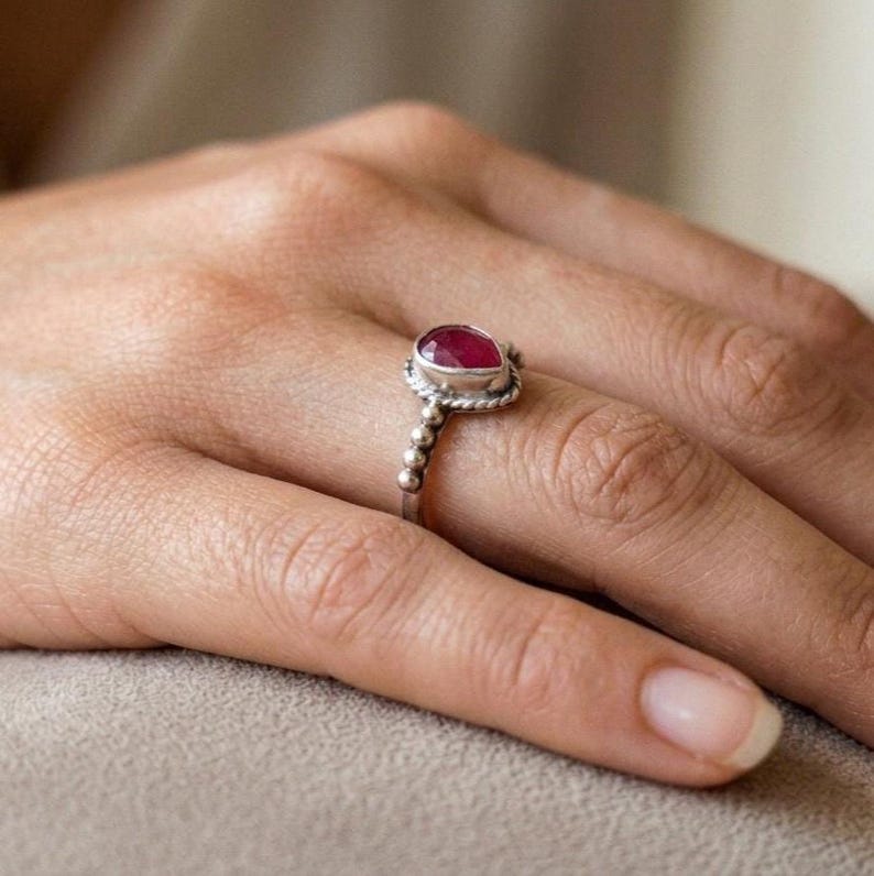 Op de afbeelding: Een zilveren ring met een peervormige, donkerroze edelsteen in een zetting. De ringband is opgebouwd uit een reeks kleine, bolvormige metalen kralen, die uitlopen in een gedraaid draaddetail rondom de centrale zetting van de edelsteen.