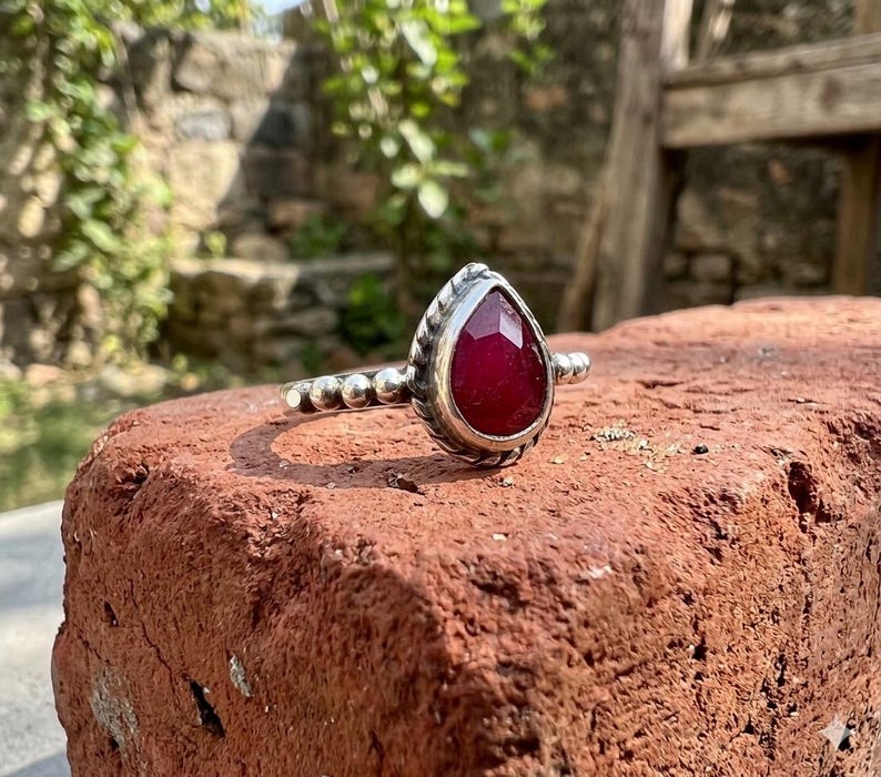 Op de afbeelding: Een zilveren ring met een peervormige, gefacetteerde dieprode edelsteen in een textuurrijke metalen zetting. De ringband bestaat uit een reeks kleine, uniforme zilveren bolletjes die vanaf de centrale zetting uitlopen in een kralenpatroon.