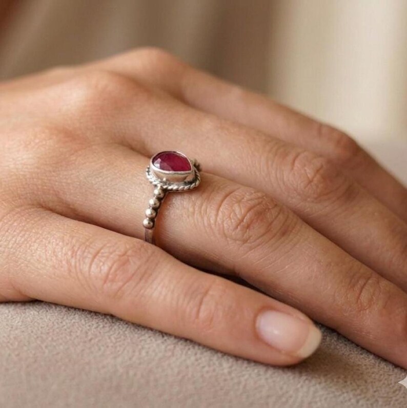Op de afbeelding: Een zilveren ring met een peervormige, diep roze-rode edelsteen in een zetting. De ringband heeft een getextureerd, kraalachtig ontwerp dat om de vinger loopt en zorgt voor een gedetailleerde metalen afwerking rond de steen.