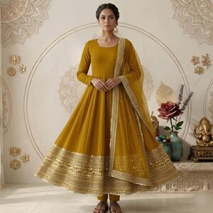 May include: Mustard yellow Anarkali suit with gold embellishments. The long-sleeved dress features a flared skirt and a matching sheer dupatta with gold trim. The outfit includes matching pants and is suitable for festive occasions.