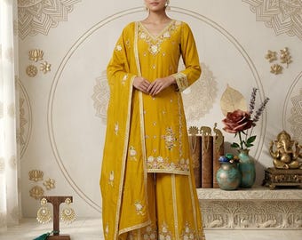 May include: A mustard yellow three-piece ensemble consisting of a long-sleeved tunic, wide-leg trousers, and a matching dupatta. The garments feature intricate white and multi-coloured floral embroidery, delicate mirror work, and scalloped borders.