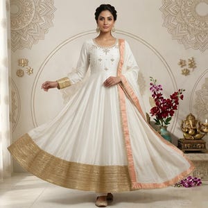 May include: A white, long-sleeved dress with gold trim and embroidery. The dress features a flowing skirt and a matching scarf with a peach-colored border. The dress is embellished with gold accents and is suitable for special occasions.
