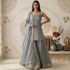 Puede incluir: Conjunto salwar kameez azul claro con bordados dorados. La parte superior sin mangas y la falda a juego se complementan con una dupatta transparente. El atuendo lo lleva una persona en una habitaci&oacute;n con decoraci&oacute;n mural.