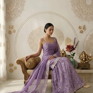 May include: A lavender-colored, embroidered Indian Sharara suit with a matching dupatta. The outfit features delicate silver embellishments and is worn with gold-colored footwear. The background includes decorative wall art and a seated figure of Ganesha.
