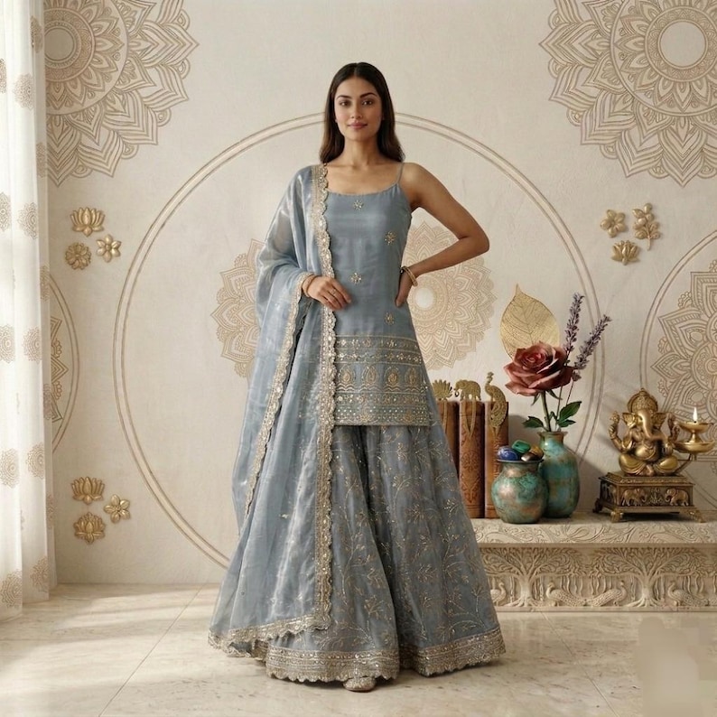 Puede incluir: Conjunto salwar kameez azul claro con bordados dorados. La parte superior sin mangas y la falda a juego se complementan con una dupatta transparente. El atuendo lo lleva una persona en una habitaci&oacute;n con decoraci&oacute;n mural.