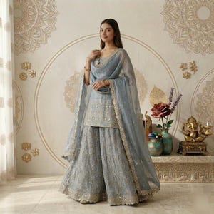 Puede incluir: Un conjunto salwar kameez indio bordado en azul claro. El atuendo incluye una t&uacute;nica, pantalones de pernera ancha y una bufanda transparente. El atuendo est&aacute; adornado con bordados y ribetes dorados. El fondo presenta arte mural decorativo y una peque&ntilde;a estatua.