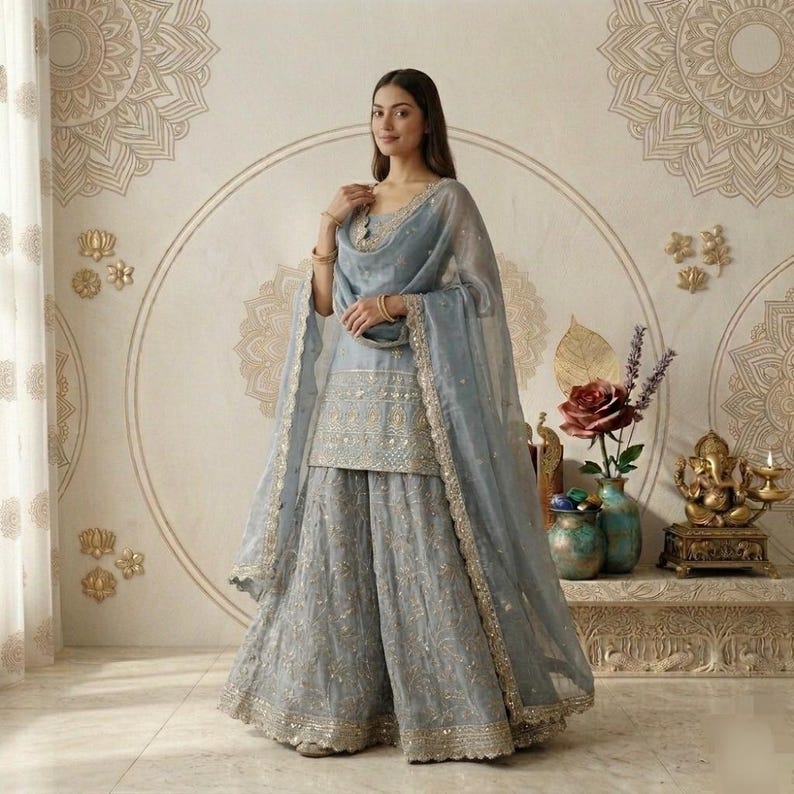 Puede incluir: Un conjunto salwar kameez indio bordado en azul claro. El atuendo incluye una t&uacute;nica, pantalones de pernera ancha y una bufanda transparente. El atuendo est&aacute; adornado con bordados y ribetes dorados. El fondo presenta arte mural decorativo y una peque&ntilde;a estatua.