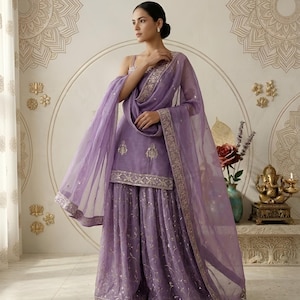 May include: A lavender Sharara suit with intricate embroidery and a matching dupatta. The ensemble features a sleeveless top and wide-legged pants, all adorned with delicate embellishments. The sheer dupatta has a gold border.