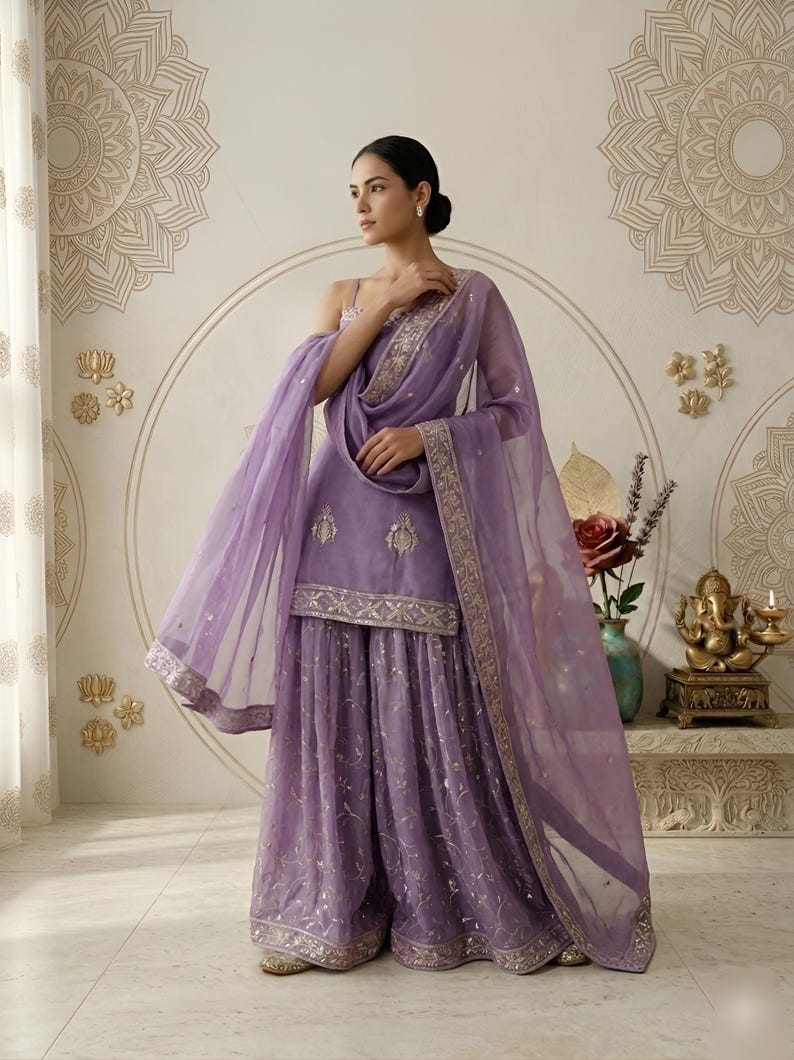 May include: A lavender Sharara suit with intricate embroidery and a matching dupatta. The ensemble features a sleeveless top and wide-legged pants, all adorned with delicate embellishments. The sheer dupatta has a gold border.