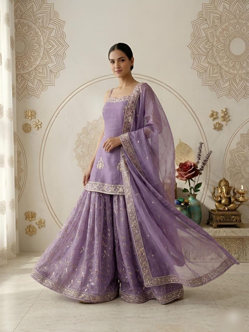 May include: A lavender-colored, formal ensemble featuring a sleeveless top and wide-leg pants. The outfit includes a matching sheer scarf with decorative trim. The ensemble is embellished with gold accents and embroidery, creating an elegant look.