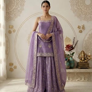 May include: A lavender-colored, three-piece Indian suit with a matching dupatta. The suit features intricate gold embroidery along the edges and on the fabric. The outfit includes a sleeveless top and wide-legged pants. The background has decorative wall art.