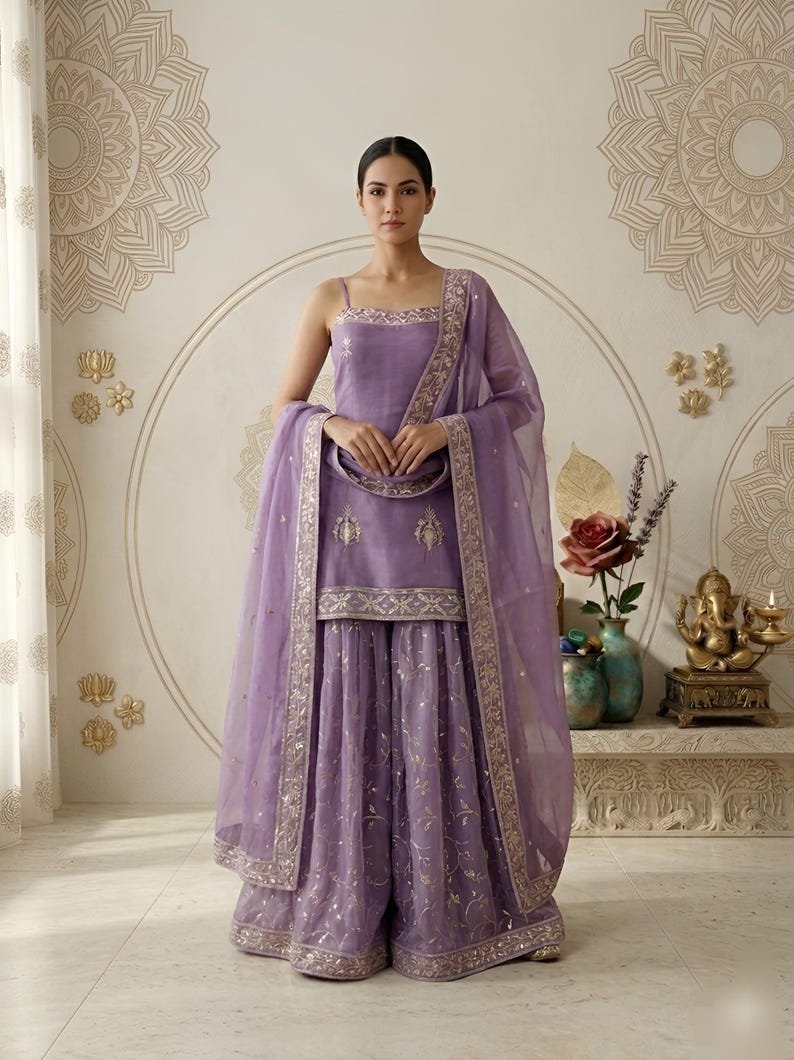 May include: A lavender-colored, three-piece Indian suit with a matching dupatta. The suit features intricate gold embroidery along the edges and on the fabric. The outfit includes a sleeveless top and wide-legged pants. The background has decorative wall art.